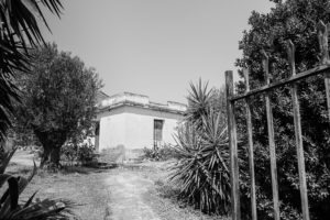 A view of a small white building from the opening gate on the property. This image is in black and white.