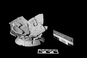 A very broken krater (mixing bowl) pictured on the left. The bowl is in pieces, with the remaining scraps of the structure laying on top of the base. A Kodak color control patch strip lies on the right. The image is in black and white.