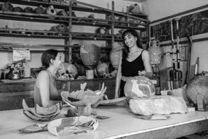 Two woman smile and talk with one another over a table of roman clay artifacts. A large shelf sits behind them full of more roman pottery and clay. This image is in black and white.
